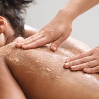 African american relaxed lady having skin scrubbing procedure at spa salon, closeup. Spa therapist applying exfoliating body mask on sleeping black woman back at luxury spa, body care concept